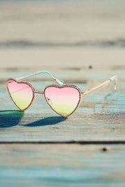 Heart Shaped Rainbow Lenses Lenses with pink rhinestones . Frame is shiny gold and they are sitting on a beachside table with beautiful sand underneath the table. Ready for the nights festivities to begin. 